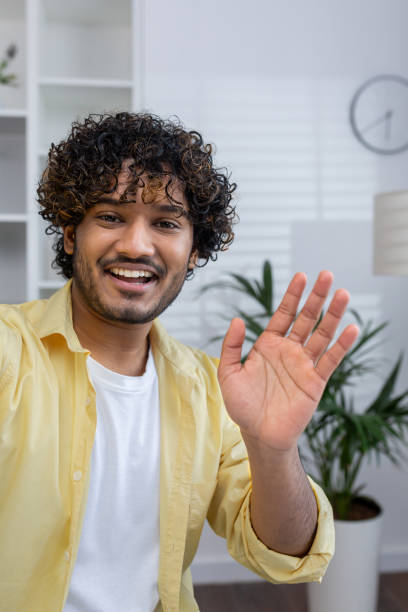 istock Smiling young man waving hello while video chatting in a modern home interior setting 2162595080
