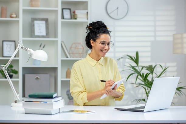 istock Hispanic woman working from home office taking notes while using a laptop in a bright modern workspace 2162596031