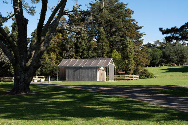 A rustic wooden shed sits in a sunlit park, surrounded by lush greenery and tall trees.
