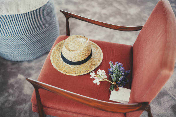 A straw hat with a black band, a small bouquet of purple and white flowers, and a blank envelope rest on a red upholstered chair. A blue-striped pouf is partially visible aloof on a carpeted floor