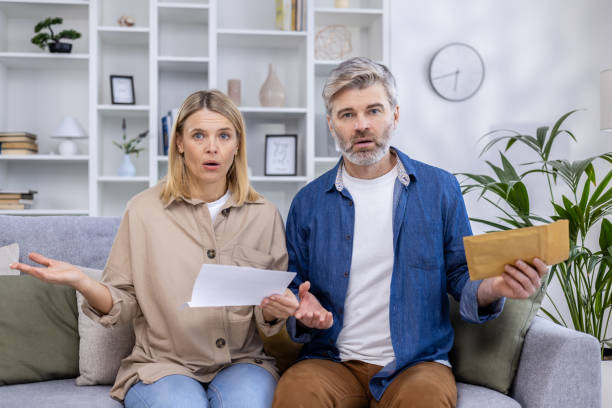 Surprised couple looking at bills and documents on their couch in living room Surprised couple sitting on their couch, holding bills and documents in their living room, shocked by unexpected expenses or financial issues. unusual couple stock pictures, royalty-free photos & images