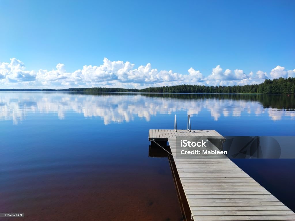 Deck at Lake Saimaa with sky reflection Small deck at Lake Saimaa on a calm summers day. Ribbon of clouds reflecting over the mirror like lake surface. Trees line the horizon line. Blue sky and lake with hint of brown. Finland Stock Photo Deck at Lake Saimaa with sky reflection Small deck at Lake Saimaa on a calm summers day. Ribbon of clouds reflecting over the mirror like lake surface. Trees line the horizon line. Blue sky and lake with hint of brown. Finland Stock Photo