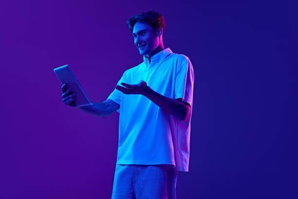 istock Emotional young man in polo shirt using tablet for online communication against gradient blue purple background in neon light 2162628710