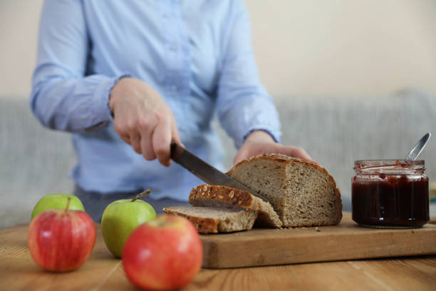 A middle-aged woman is captured up close while slicing a loaf of bread at a kitchen table. She is wearing a blue shirt, and the focus is on her hands and the bread, showcasing the detail and care involved in food preparation. The kitchen table and surrounding elements hint at a warm and inviting home environment. This image is perfect for use in cooking, home life, and culinary-themed content, emphasizing the personal touch and everyday moments of preparing meals.