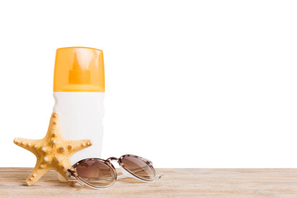 A woman wearing a sun hat applies sunscreen from a spray bottle on her hands while enjoying a sunny day at the beach.