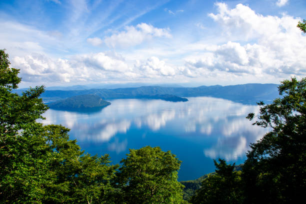A beautiful summer scenery of Lake Towada in Aomori, Japan. The clear waters reflect the vibrant blue sky, creating a picturesque landscape. This serene location is perfect for nature lovers and outdoor enthusiasts. The image captures the beauty and tranquility of Lake Towada during the summer season.