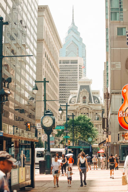 Philadelphia, Pennsylvania, July 19, 2024. Pedestrians walk along a bustling city street lined with tall buildings. In the background, a prominent clock tower and a modern skyscraper can be seen amid the urban landscape.