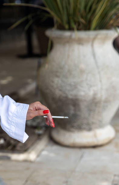Female hand with red nails and white blouse holding a cigarette on blurred background of large ceramic pot outdoors.content related to lifestyle, habits and relaxation.