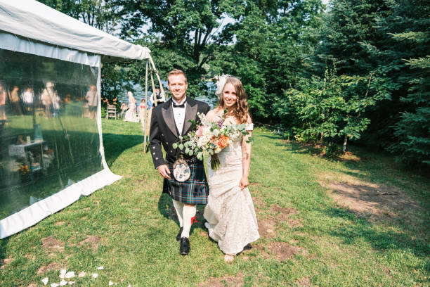 Millennial wedding ceremony outdoors in summer. One spouse is a non-binary person, dressed in a Scottish kilt with a beret, body positive bride dressed in white, maid of honour and best man on the side. Celebrant is standing. Horizontal full length outdoors shot. This was taken in Quebec, Canada.