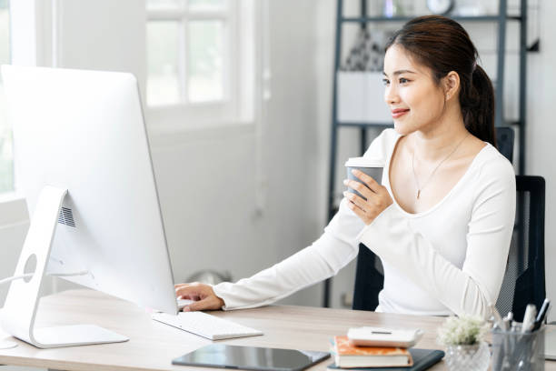 istock Beautiful young woman in casual clothing using desktop computer  and enjoying hot drink while working indoors 2162680145