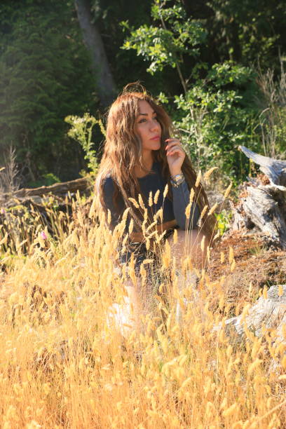 An Iranian woman sitting outdoors on a sunny Summer day. She is behind some wild ripening grass on the edge of a forest. She has long, brown, curly hair, makeup, a bracelet and a long sleeved shirt.
