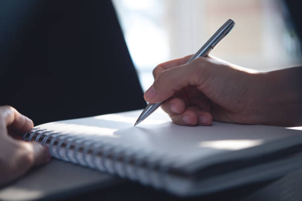 istock Close up, woman hand writing on spiral notebook wuth laptop computer on table. Female hand taking note on paper notepad, business planning, reminder, to do list, backlit 2162757339