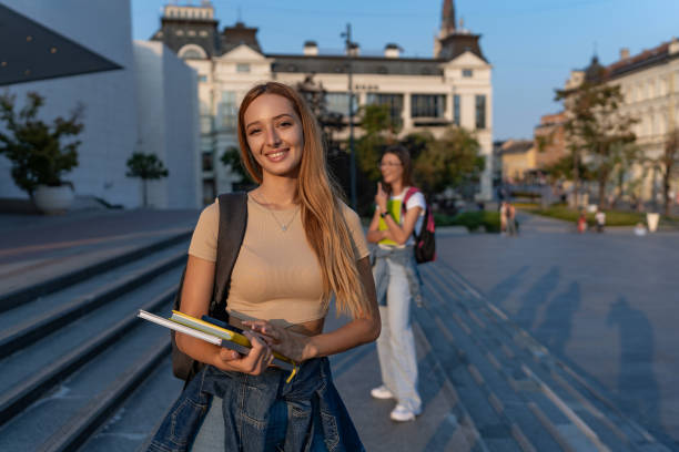 A female student in a cropped top smiling and holding books on the campus steps, with a flannel shirt tied around her waist and a pleasant urban setting in the background.