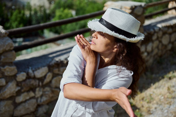 A woman wearing a white hat stands outside with arms raised, capturing a moment of tranquility and freedom in a serene natural environment.
