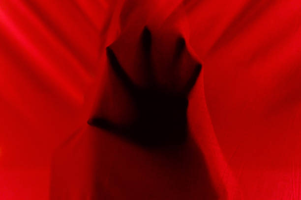 Unrecognizable mysterious person with hand hiding behind red translucent light curtains while standing on murk balcony in apartment at dark night time