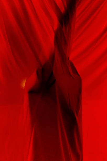 Unrecognizable mysterious person hiding behind red translucent light curtains while standing on murk balcony in apartment at dark night time