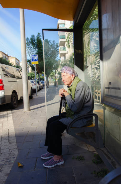 An elderly woman at a bus stop seems to be either napping or soaking in the sun's gentle rays.