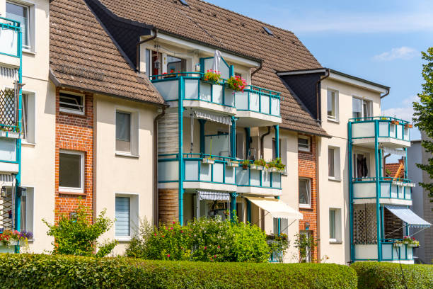 A modern residential complex built for social housing. The buildings have multiple stories and feature a varied facade of red bricks and white plaster surfaces. Notable are the blue balcony railings and window frames, adding colorful accents. Each balcony is adorned with flowers, giving the buildings an inviting and vibrant character. Well-maintained green hedges and bushes surround the complex, which is captured under a clear blue sky on a sunny day