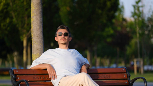 A handsome, young, dark-haired man striking cool poses while sitting happily on a bench in a tree-filled park on a summer day