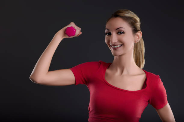 Confident young woman in athletic gear smiles while flexing her bicep with a pink dumbbell, showing determination and motivation in her fitness goals