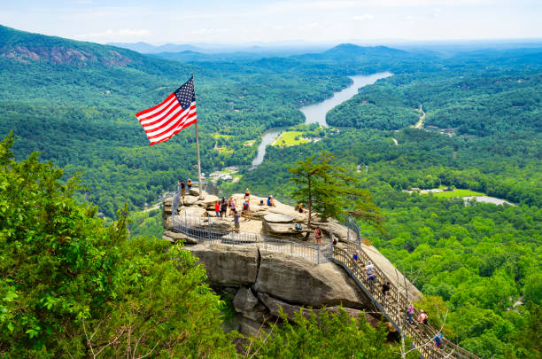 Chimney Rock State Park, North Carolina, United States - May 22, 2024: Tourists at the top of Chimney Rock,, with a waving American flag, and Lake Lure in the background,