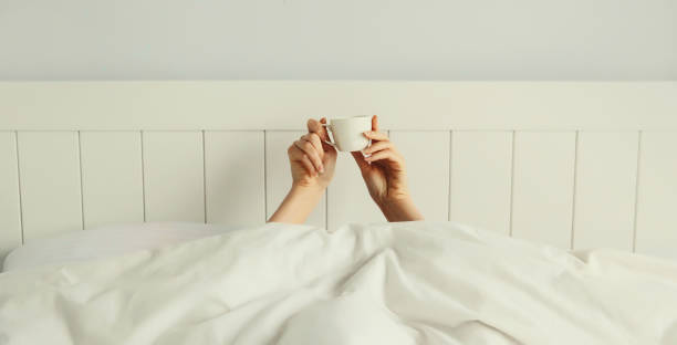 istock Cheerful lazy woman waking up after sleeping lying in bed holding cup in her hands asks for coffee 2162870058