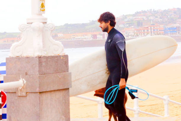 Side view of one surfer wearing wetsuit, carrying his surfboard on the sidewalk by San Lorenzo beach, Gijón, Asturias , Spain.