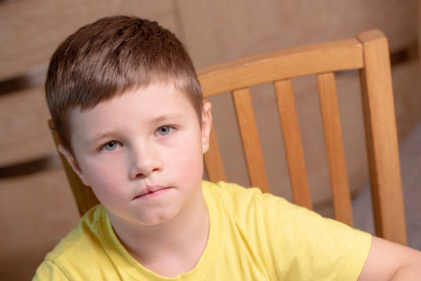 With a stern look, a boy in a yellow T-shirt sits on a wooden chair, glaring at the camera and showing clear signs of irritation