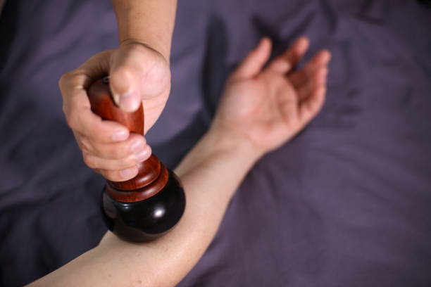 A middle-aged Caucasian male therapist in a white uniform uses a kamni bowl for a healing session with a relaxed Hispanic female client lying on a treatment table in a calming, well-lit setting.