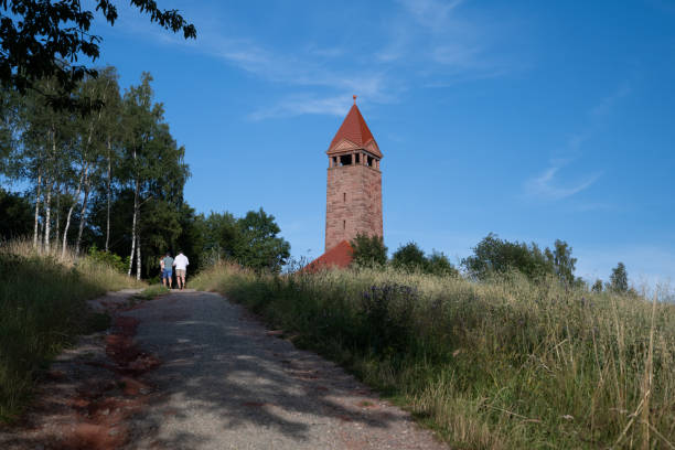 An observation tower near Nowa Ruda in southwest Poland. Unrecognizable people are seen walking up a hill towards the tower, surrounded by tall grass and trees under a clear blue sky.