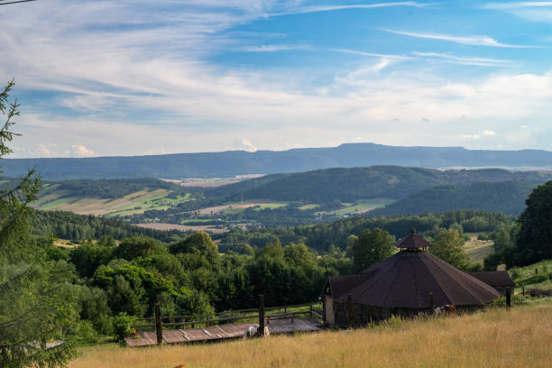 This picturesque landscape captures the serene beauty of the southwest Polish countryside. A quaint octagonal building with a red roof sits prominently in the foreground, surrounded by lush green hills and valleys. The rolling landscape extends into the distance, featuring a mix of dense forests and open fields under a blue sky with wispy clouds.