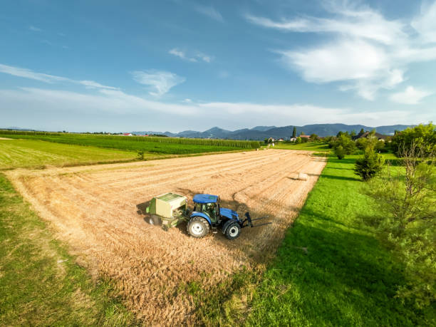 A drone point of view, looking down at a farmer driving a blue tractor, that is towing a green hay baler. Lines of dried cut grass are being fed into the baler and being made into large round bales of hay, that will be used as winter feed for livestock. There are beautiful patterns of harvesting that can only be seen from above.