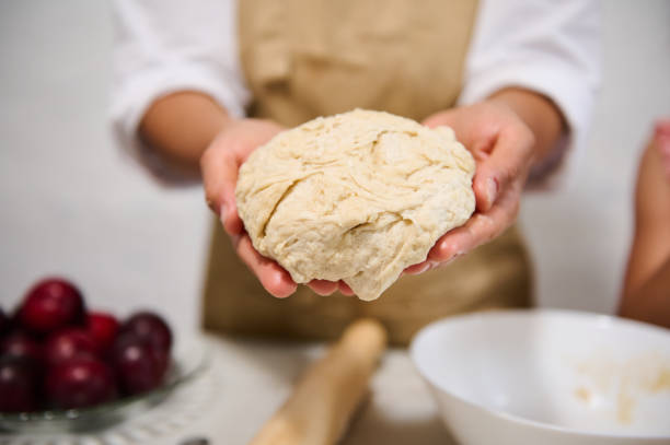 Close-up of a person holding raw dough in their hands, preparing for baking. Fresh ingredients like plums and flour can be seen in the background.