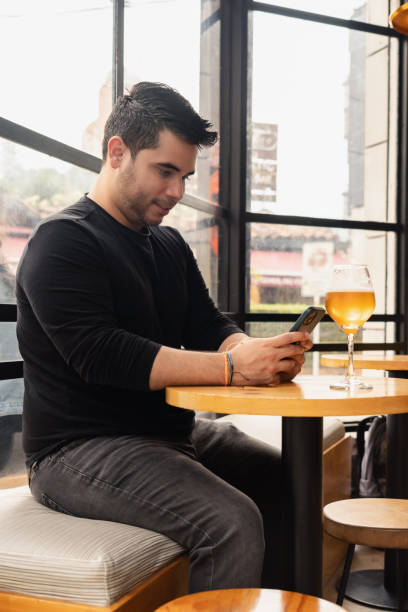 istock Young Latino man using cell phone in a bar, drinking craft beer at a table. 2162943884