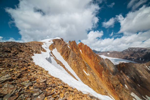 istock Snow-capped mountain ridge with cornice and red sharp rocks on top in sunlight. Snow cap and shiny pointy peak of gold color. Snow dome and colorful rocky peaked top. Sunny cloudy high mountains view. 2162946189