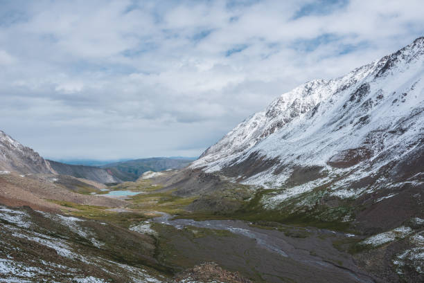 istock Dramatic landscape with river meanders and mountain lake in green stone valley among snow covered hills in rainy cnangeable weather. Freshly fallen snow in high mountains under clouds in blue sky. 2162946191