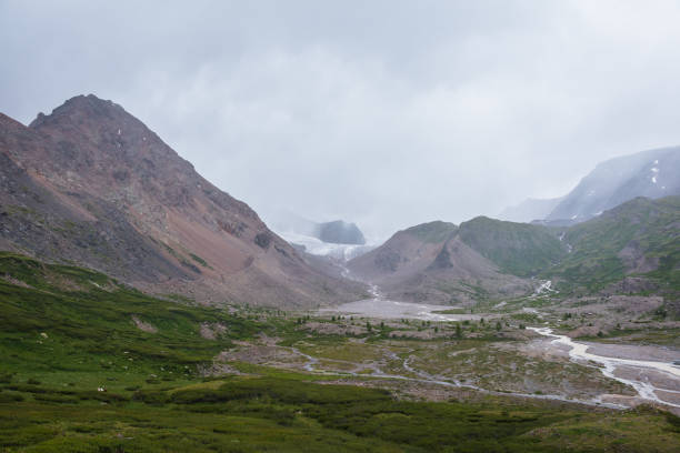 istock Mountain river flows from big glacier among large rocky snowy mountains in low clouds into green alpine valley with lush flora under rainy gray cloudy sky. River floods between small grassy islands. 2162946193