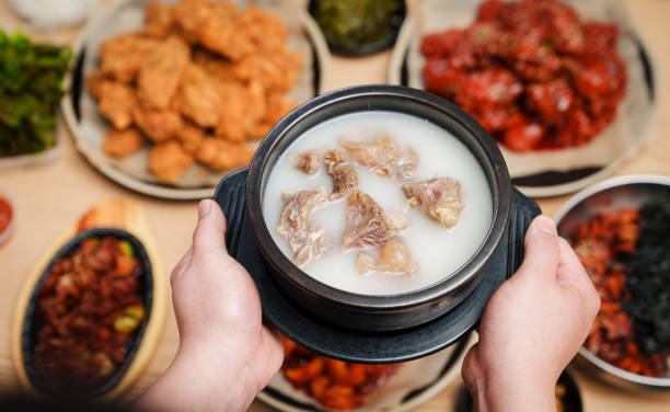 Human hands holding bowl of ox knee soup with Korean food in background.