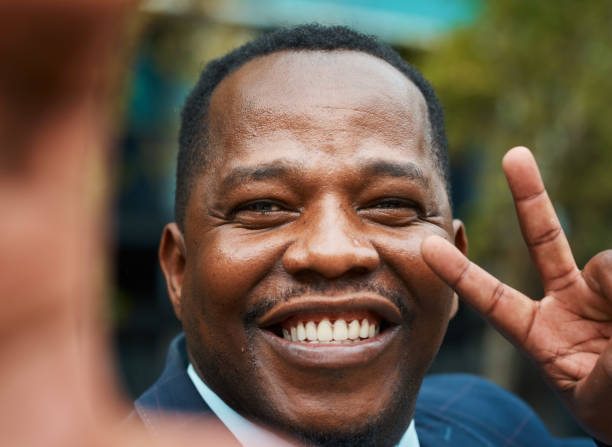 Close-up portrait of a cheerful black businessman making a peace sign, capturing a moment of positivity and confidence in a bustling city environment.