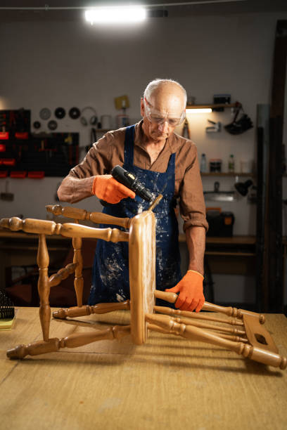 istock Elderly man in a workshop using a drill with a special attachment to remove old varnish from a wooden chair. He is wearing protective goggles and orange gloves. 2162984355