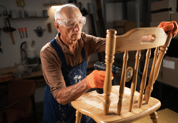 istock An elderly man in a workshop uses a drill with a special attachment to remove old varnish from a wooden chair. He is wearing protective glasses and orange gloves. 2162984356