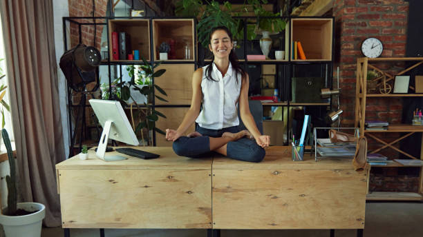 istock Woman, smiling office worker sitting on desk surrounded by computer and office supplies, doing calming meditation in yoga lotus pose 2162985509