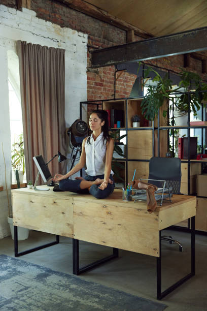 istock Woman, office worker sitting on desk surrounded by computer and office supplies, doing calming meditation in yoga lotus pose 2162985529