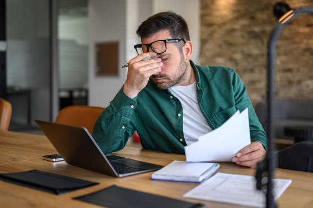 Anxious male financial advisor rubbing eyes while working over laptop and analyzing reports at desk