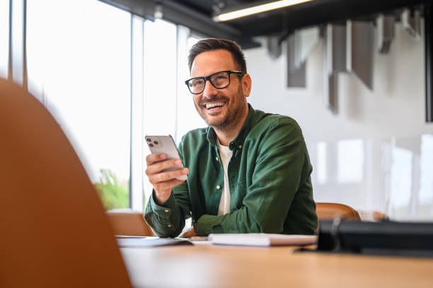 istock Cheerful handsome businessman in eyeglasses looking away while using mobile phone at desk in office 2162997970