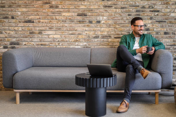 istock Positive male entrepreneur with coffee cup looking away thoughtfully while relaxing on sofa in office 2162997977