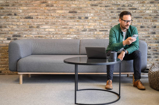 istock Confident freelancer with laptop and coffee cup on table texting over smart phone on sofa in office 2162997982