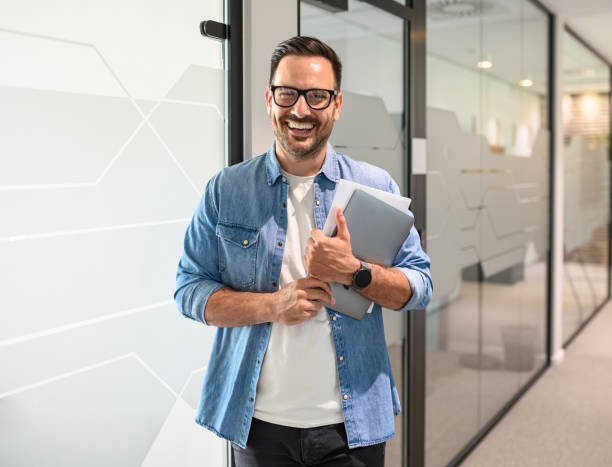 istock Confident male professional with computer and document smiling and standing by glass wall in office 2162997995