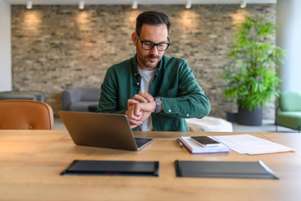 Male project manager checking time over wristwatch while working over computer at desk in office