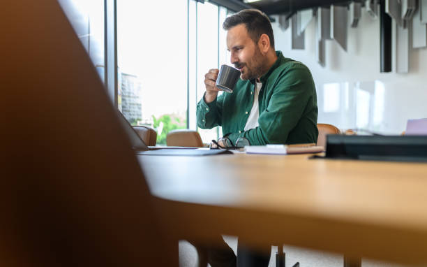 istock Male project manager drinking coffee while relaxing at wooden desk in modern office during breaks 2162998050